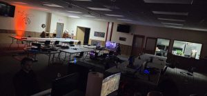 Large dim room set up for a LAN party with long tables, multiple computer workstations, and people gaming.